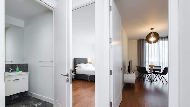 A modern hotel room with a bathroom, a bedroom with a bed and bedside table, and a dining area with a table and chairs. - Fernbank by Dublin At Home (Dublin 14)