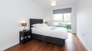 A modern bedroom with a large bed, a nightstand, a lamp, and a window with a view of greenery. - Fernbank by Dublin At Home (Dublin 14)
