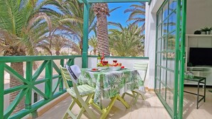 A balcony with a table set for dining, surrounded by palm trees and a TV in the background. - Apartment PALM GARDEN - Costa Calma - Fiber Optic Internet - Pool (Costa Calma)