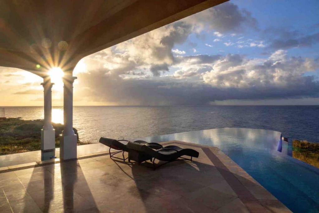 An infinity pool with lounge chairs under a covered area, overlooking the sea.