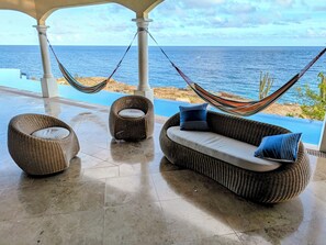A hammock, two wicker chairs, and a sofa with cushions by a poolside with a view of the sea.