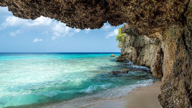 A beach with clear turquoise water, a rocky cliff, and a cave opening. - Award-Winning Luxury Beach-Side Villa - Wrap-around Infinity Pool -Pickleball (Sabana Westpunt)
