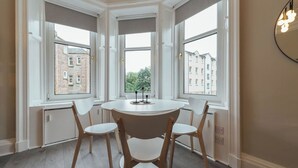 Ein Zimmer mit einem runden Tisch, vier Stühlen, großen Fenstern mit Blick auf Gebäude und einer Wandleuchte.