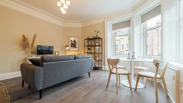 Ein modernes Wohnzimmer mit einem grauen Sofa, Holzstühlen, einem Fernseher, einem Bücherregal und einem Fenster mit Blick auf Gebäude.