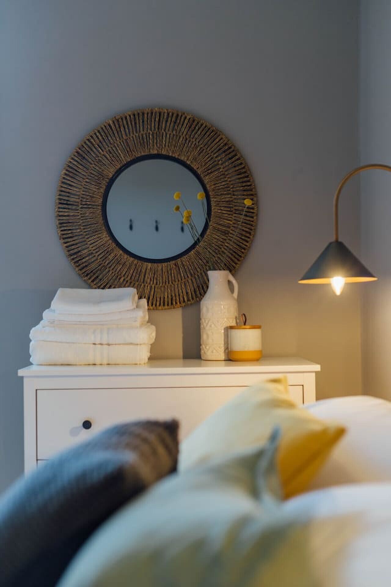 Et pænt møbleret soveværelse med et rundt spejl, en sengetårnlampe, en stak foldede håndklæder, en hvid vase og et lille lys på et hvidt kommode.
