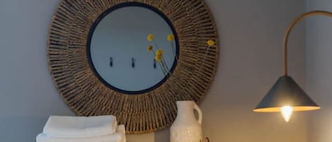 Et pænt møbleret soveværelse med et rundt spejl, en sengetårnlampe, en stak foldede håndklæder, en hvid vase og et lille lys på et hvidt kommode.