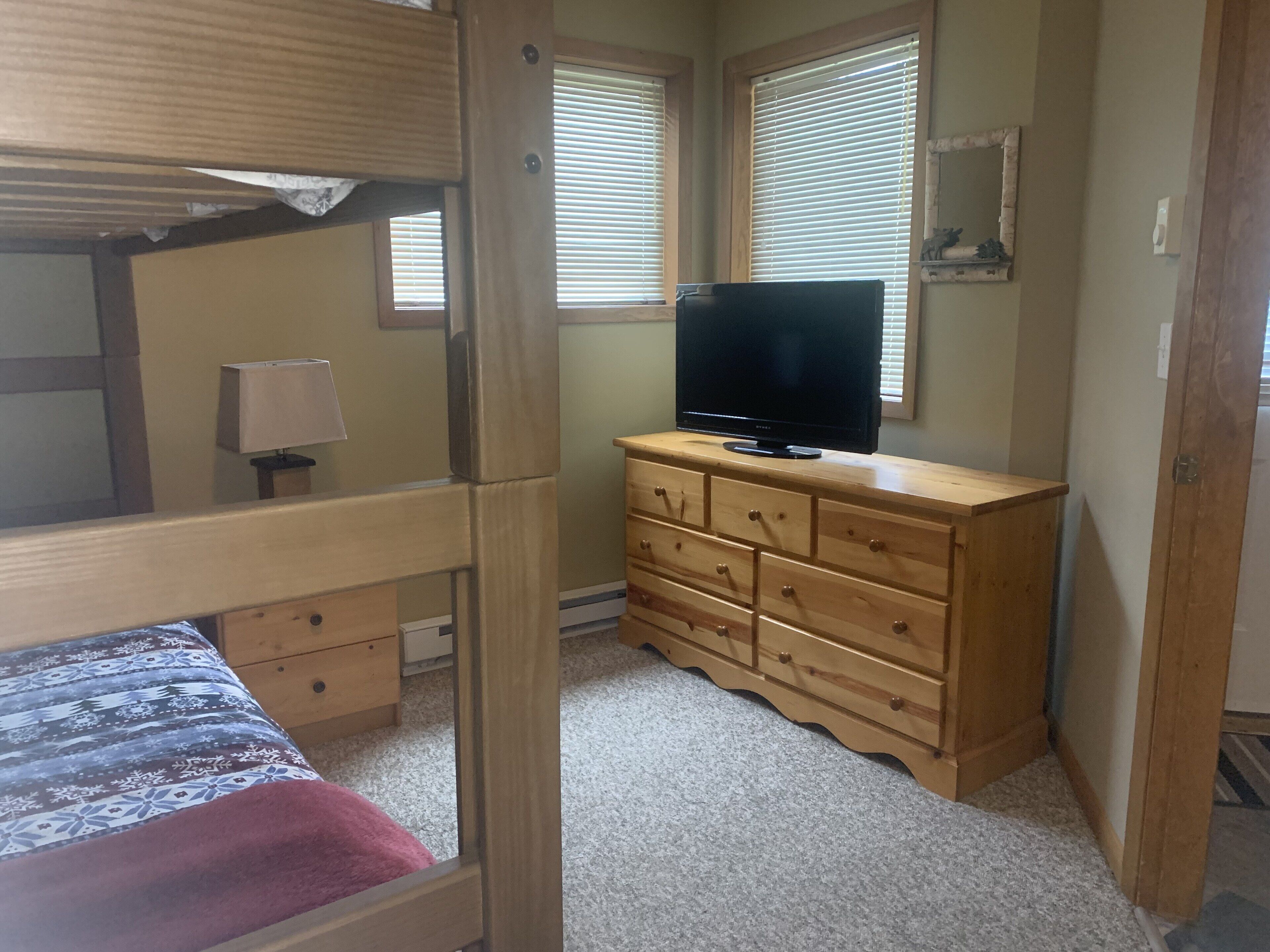 Quarto com beliche, cómoda de madeira com gavetas, televisão em cima e janela com persianas.