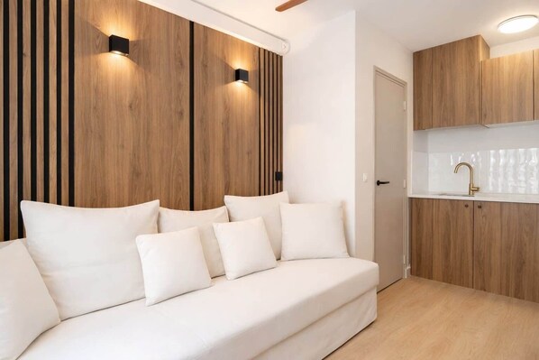 A modern living room with a sofa, a kitchen area with wooden cabinets, and a wall-mounted light fixture.