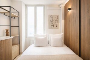 A bedroom with a bed, a wooden wardrobe, a white shelf unit, and a window.