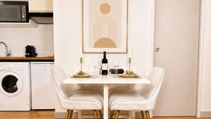 Un'area pranzo con un tavolo bianco, due sedie bianche a gambe dorate, una bottiglia di vino e due bicchieri sul tavolo.