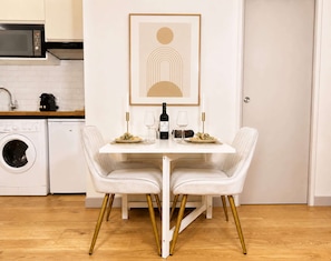 A dining area with a white table, two white chairs with gold legs, a bottle of wine, and two glasses on the table.