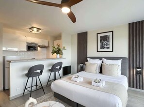 A modern hotel room with a bed, bedside tables, a desk, two chairs, a kitchen area, and a ceiling fan.