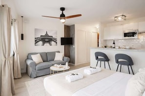 A modern hotel room with a bed, a sofa, a small table, a TV, a ceiling fan, and a kitchenette.