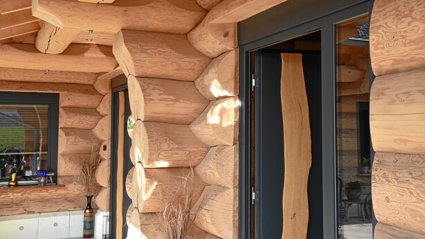 Interior de una cabaña de troncos con techo de madera, ventanas amplias y una puerta que conduce a otra habitación.