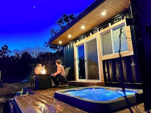 A hot tub with two people sitting on chairs, a wooden deck, and a glass door leading to a covered area. - Private Glamping Container • Hot Tub Escape • Creek Access (Monument)