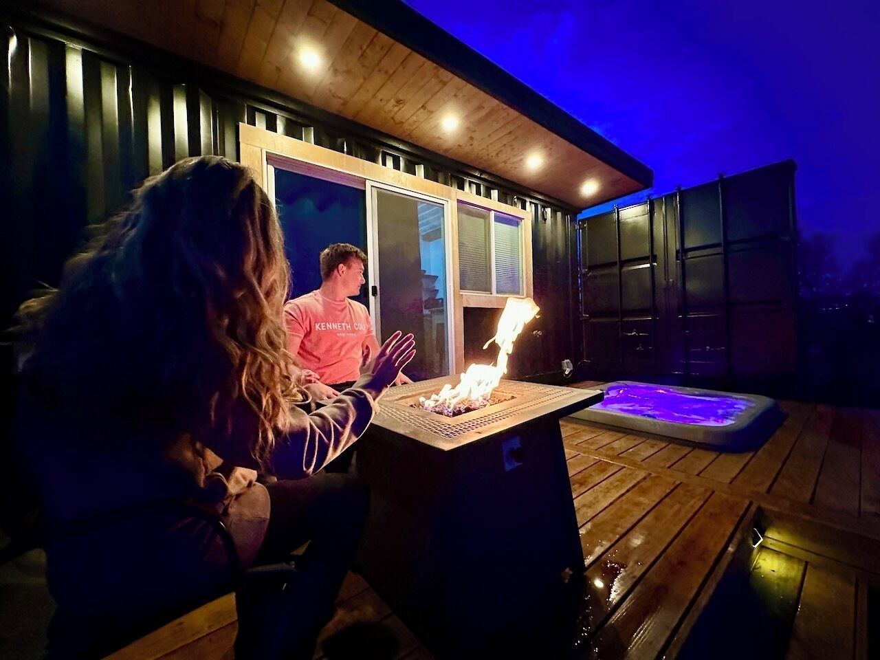 Iemand zit aan een tafel met een vuurplaats, in een buitengebied met een houten terras en een glazen deur die naar een gebouw leidt.