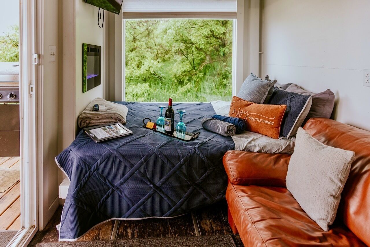 A bedroom with a bed, a window with a view of greenery, a small table with a bottle and glasses, and a leather sofa.