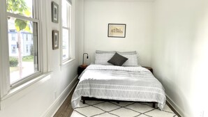 Een slaapkamer met een bed, een nachtkastje, een lijst met een foto aan de muur en een raam met uitzicht op gebouwen.