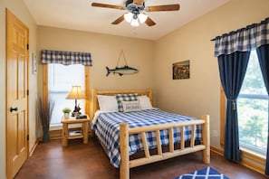 Een slaapkamer met een houten bed, een plafondventilator, een raam met gordijnen, een nachtkastje met een lamp en een visdecoratie aan de muur.