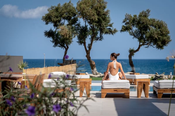 A woman sitting on a lounge chair by a pool with a view of the sea and trees.