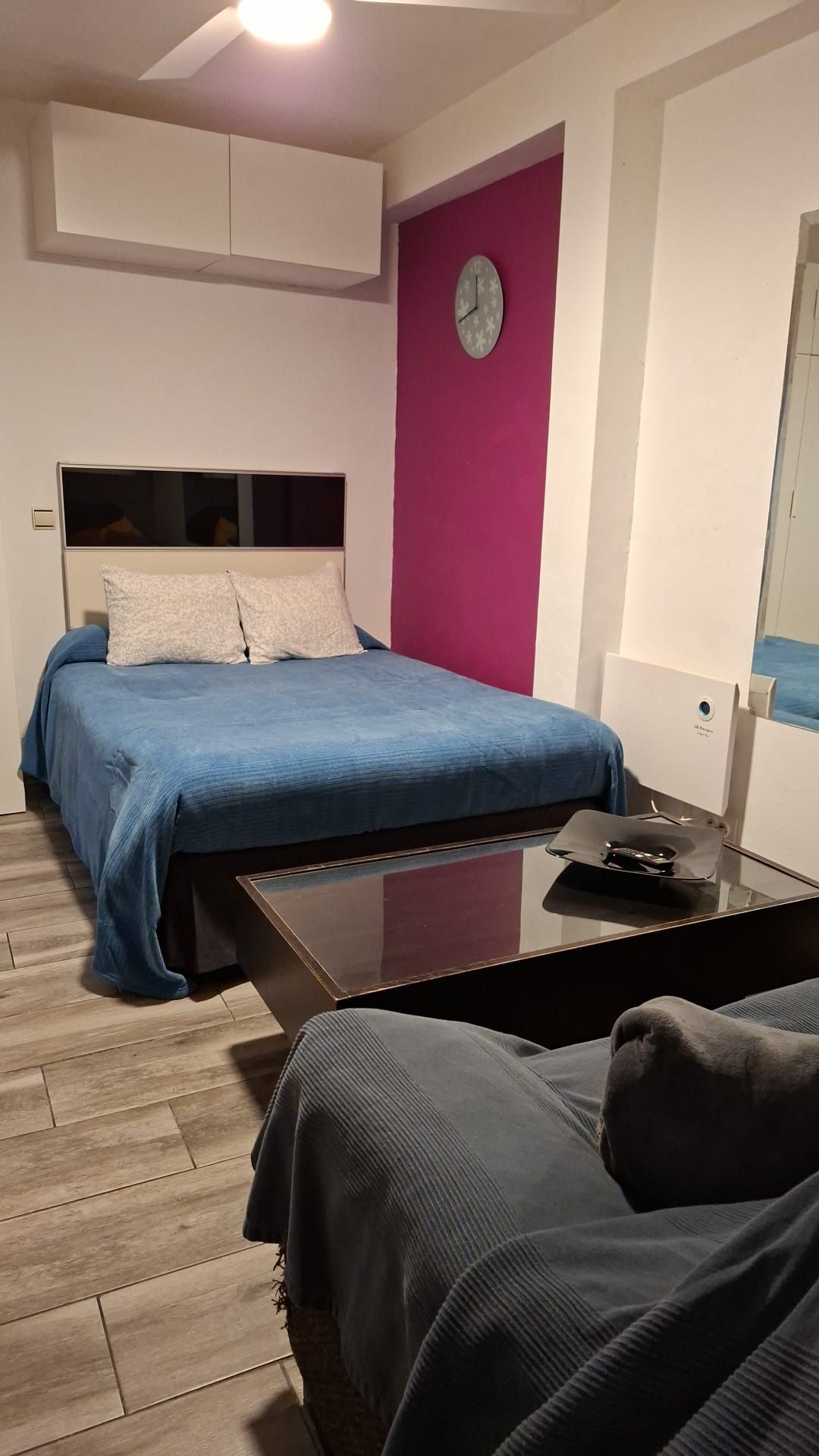 A bedroom with a bed, a bedside table, a chair, a clock on the wall, and a window.