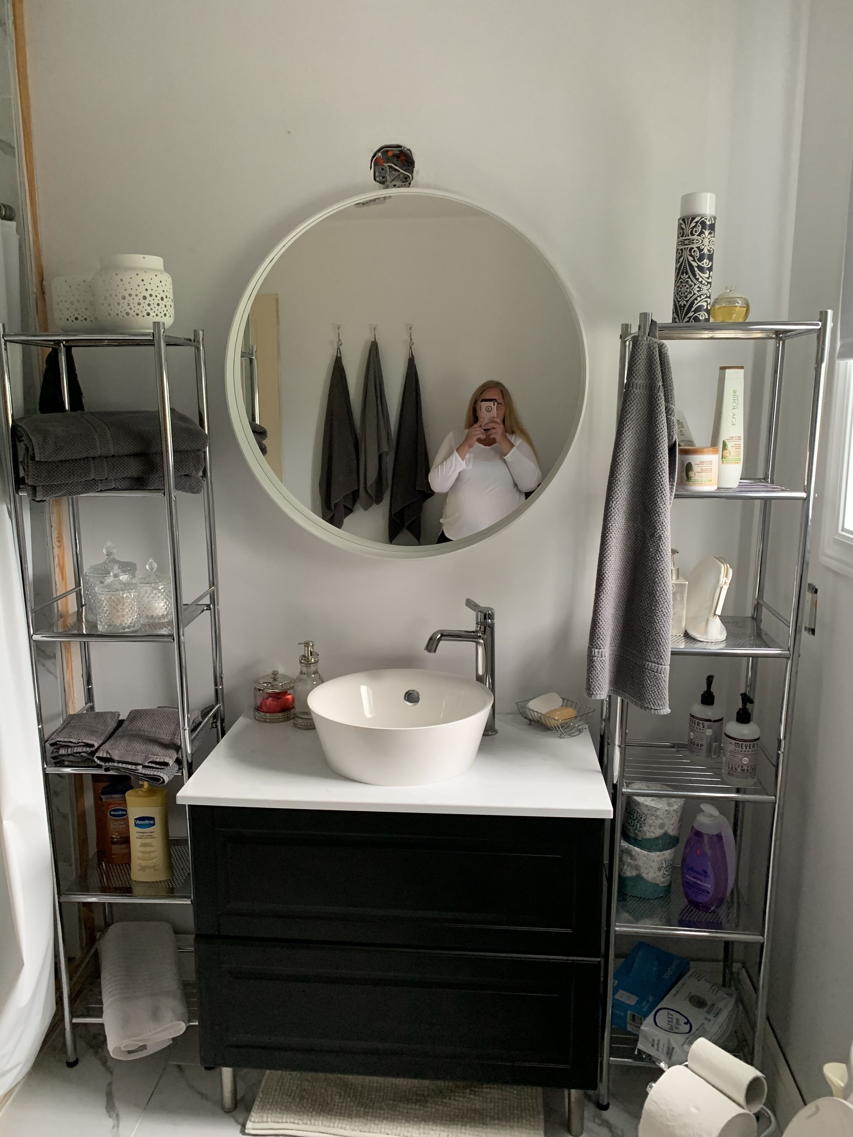 Une salle de bain avec un miroir rond, un lavabo blanc et un meuble-lavabo noir. On y trouve des étagères avec des serviettes et des articles de toilette.