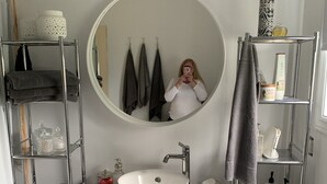Ein Badezimmer mit einem runden Spiegel, einem weißen Waschbecken und einer schwarzen Kommode. Es gibt Regale mit Handtüchern und Pflegeprodukten.