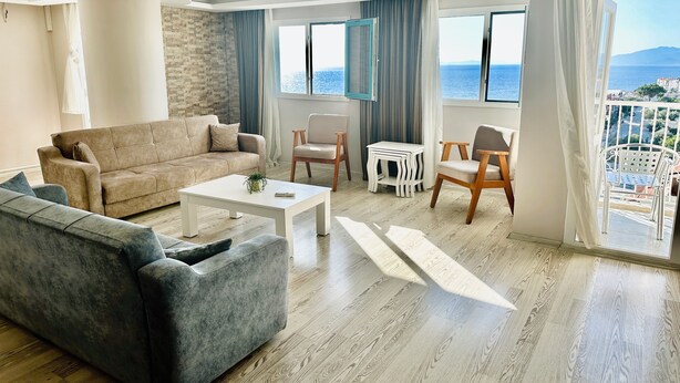 A modern living room with a grey sofa, a white coffee table, and a wooden floor. There are two windows with blue curtains, and a balcony with outdoor furniture is visible through the window. - Silver Tower Family Apart Otel (Kusadasi)