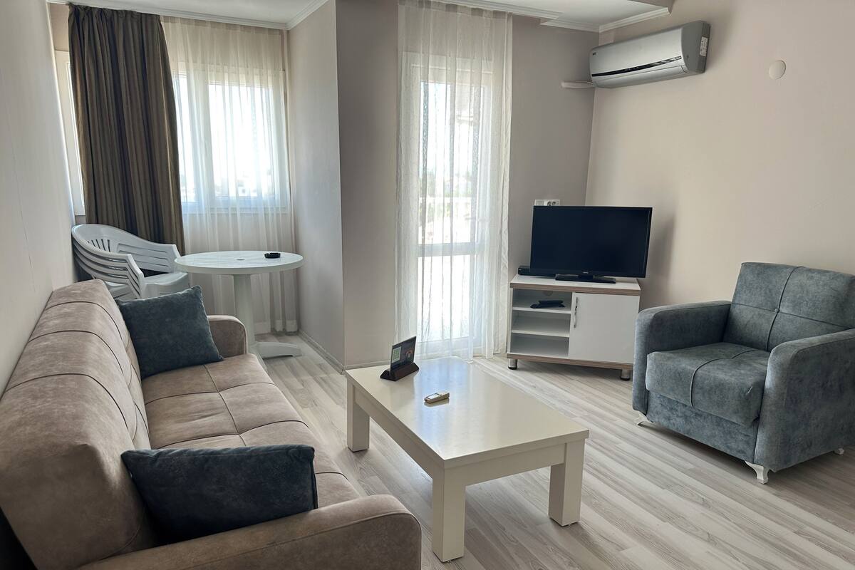 A modern living room with a beige sofa, a grey armchair, a white coffee table, a flat-screen TV on a white stand, and a ceiling-mounted air conditioner.