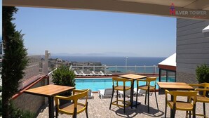 A terrace with a pool, yellow chairs, and a view of the sea and distant buildings. - Silver Tower Family Apart Otel (Kusadasi)