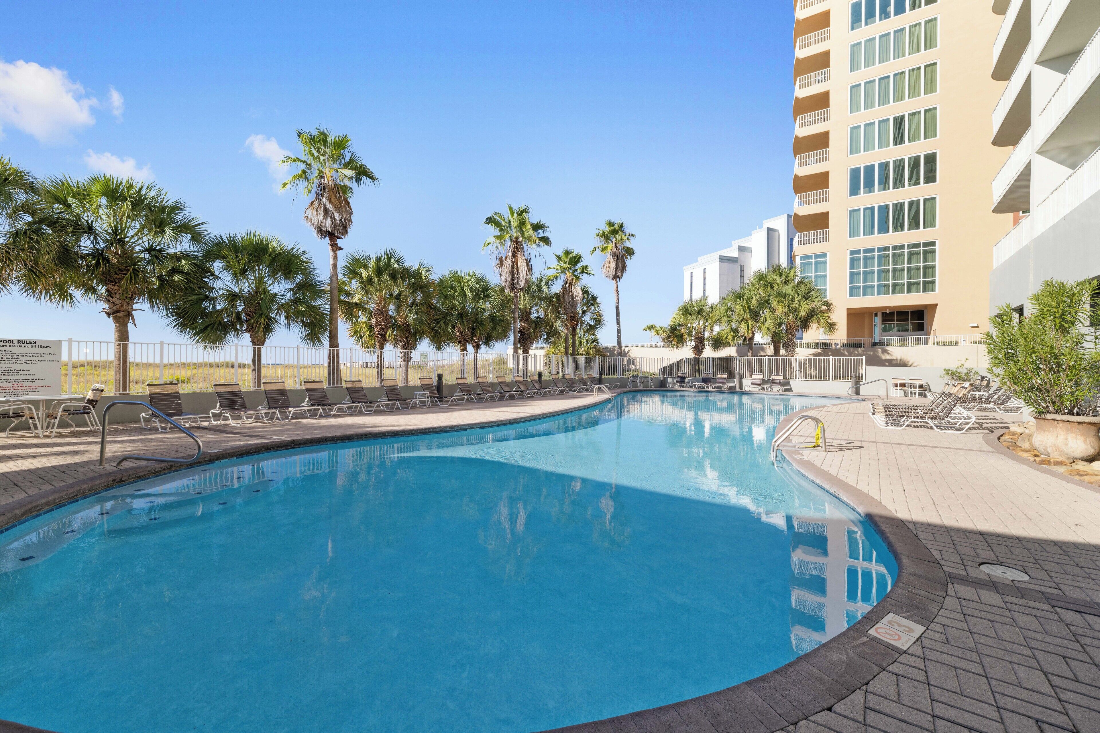 A swimming pool with lounge chairs and palm trees, surrounded by a building with balconies.