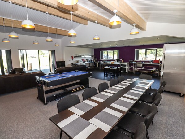 A pool table in a room with a long table and chairs, a sofa, and a kitchen area with a refrigerator.