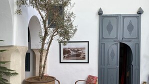 A courtyard with a large potted tree, a framed photograph, a patterned chair, and a decorative door. - Riad Magellan with swimming pool & Jaccuzzi 2 steps from Jamaa el Fna square (Marrakech)