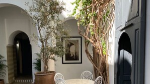 A courtyard with a tiled floor, a round table with chairs, a large potted plant, and a wall-mounted picture. - Riad Magellan with swimming pool & Jaccuzzi 2 steps from Jamaa el Fna square (Marrakech)