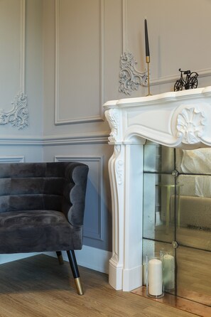 A room with a dark upholstered armchair, a white fireplace with decorative moldings, and a bicycle in the background.