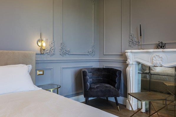 A hotel room with a bed, a chair, a small table, a fireplace, and wall-mounted sconces.