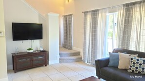 A living room with a flat-screen TV, a wooden entertainment unit, a sofa with patterned cushions, and a staircase leading to another level. - Asteria – Your perfect stay close to Theme Parks. (Kissimmee)