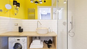 A modern bathroom with a yellow accent wall, a round sink, a glass shower enclosure, and a washing machine.