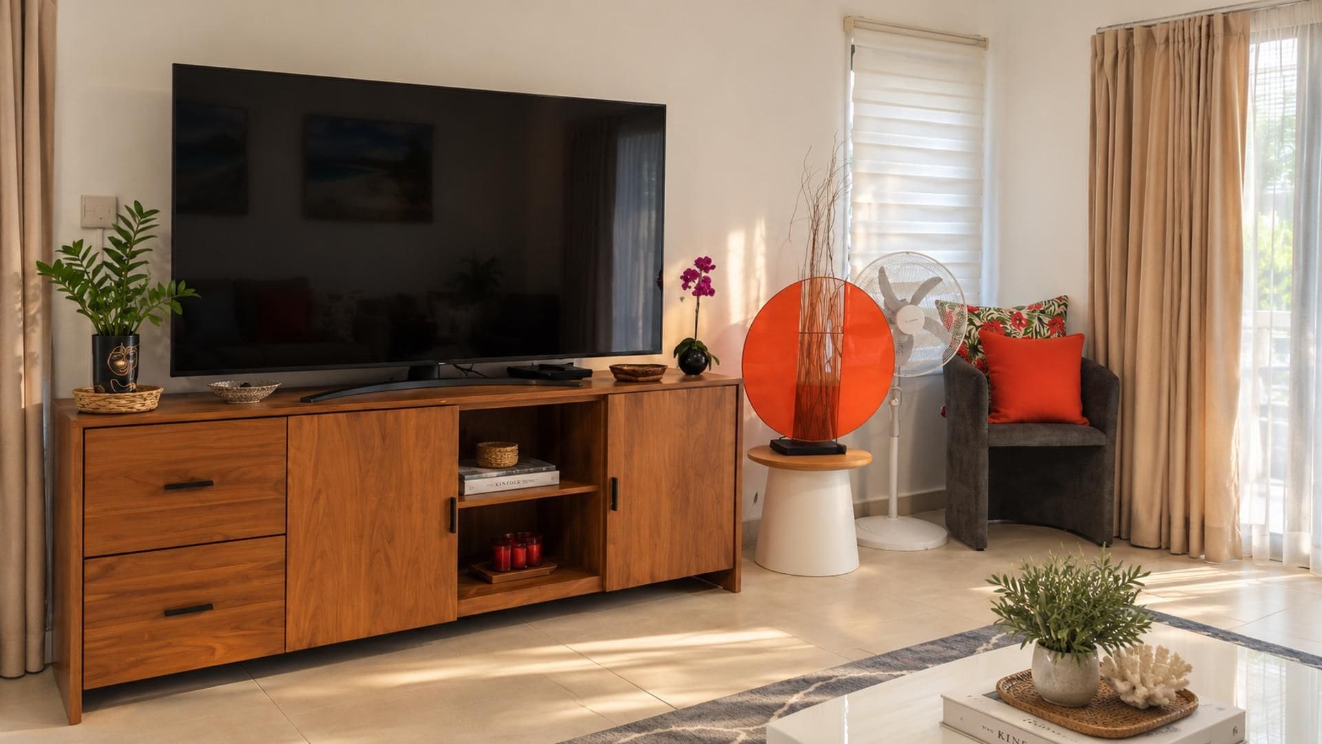 Un soggiorno moderno con una TV a schermo piatto su un mobile in legno, un elemento decorativo arancione a forma circolare, un divano grigio con un cuscino rosso e una pianta in vaso su un tavolino da caffè.