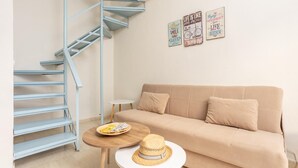 A living room with a beige sofa, a blue spiral staircase, a small round table with a straw hat, and three motivational wall art pieces. - Flora Maisonettes 1 - Two Bedroom House, Sleeps 4 (Lakka)