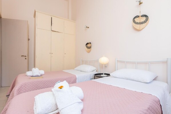 A hotel room with two beds, each with a pink quilted bedspread and white pillows. There is a wardrobe with a door, a bedside table with a lamp, and a decorative wall hanging.