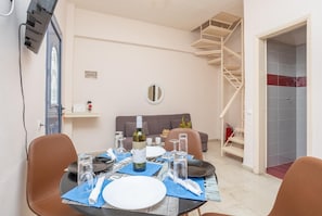 Un comedor con una mesa lista para dos personas, un sofá, una escalera en espiral y una zona de ducha con azulejos.