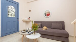 A room with a blue door, a grey sofa, a small white table with a vase of flowers, and a wall-mounted mirror.