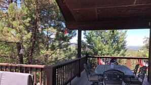 A covered deck with a wooden floor, a sofa, and a table with chairs. - BearLuckLodge! Amazing Cabin-Hot Tub, Views, Game Room/Arcade, Location, Peloton (Ruidoso)