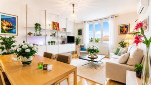 Moderni olohuone, jossa on puinen ruokapöytä, sohvia ja sohvapöytä. Huoneessa on myös ruukkukasveja, televisio ja taideteoksia seinillä.