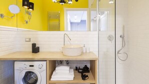 A modern bathroom with a yellow accent wall, a round sink, a built-in washing machine, and a glass shower enclosure.