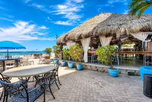 Un restaurante frente a la playa con terraza, estructuras de techo de paja y plantas en macetas.