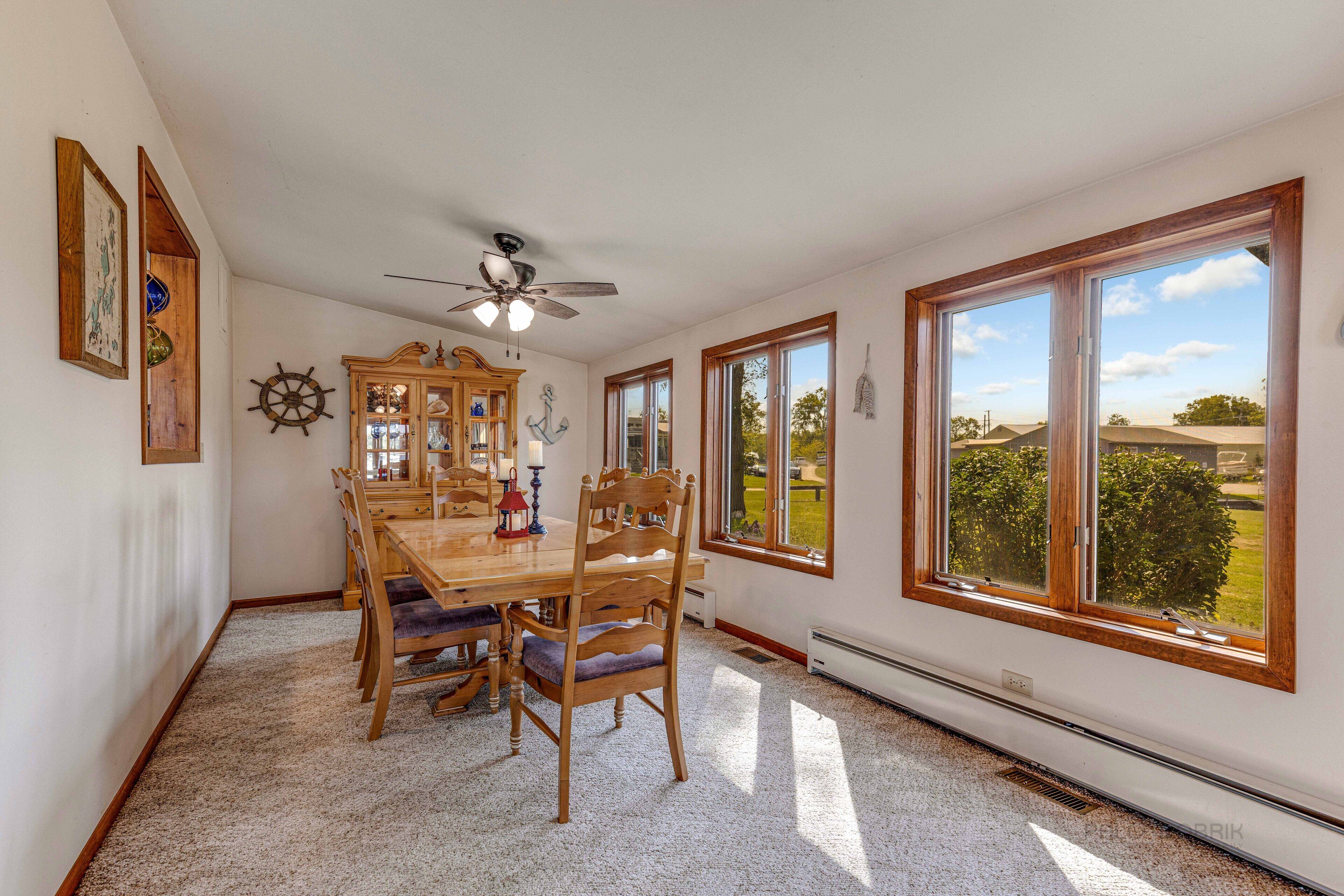 A dining room with a wooden table and chairs, a ceiling fan, and large windows with a view of the outdoors.