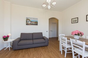 A modern living room with a grey sofa, a wooden dining table with white chairs, a chandelier, and a painting on the wall. - Costa Azzurra (Stresa)