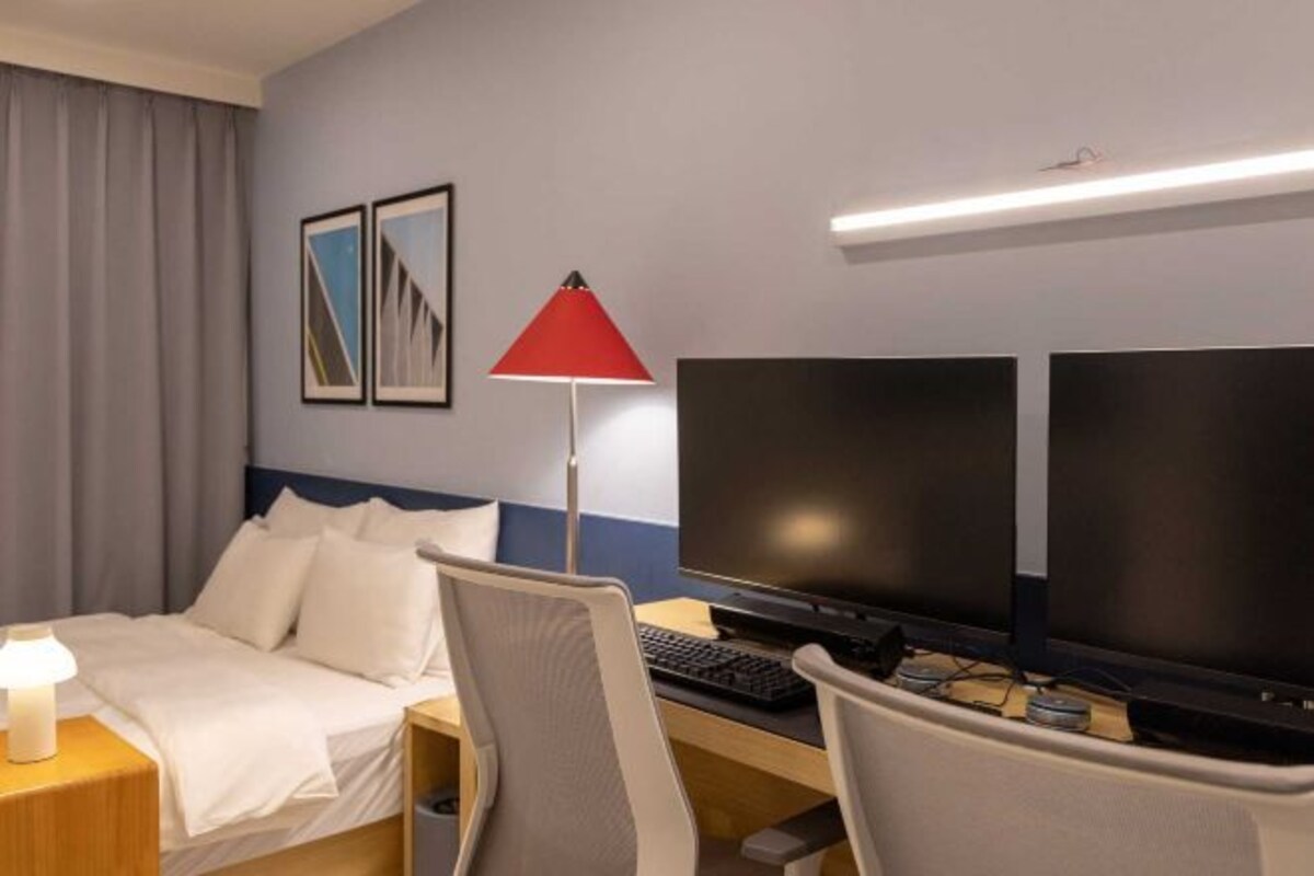 A hotel room with a bed, desk, two monitors, a lamp, and a picture on the wall.